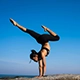 Person performing a handstand split on rocky terrain under clear blue sky, showcasing advanced yoga balance, core strength, and flexibility in a scenic outdoor setting.