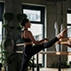 Ballet dancer practicing high leg lift at barre in sunlit studio, wearing black athletic attire—capturing grace, flexibility, and focused training in a serene, plant-filled space.