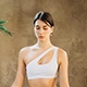 Woman meditating in white asymmetrical sports top, seated with eyes closed in serene indoor space with neutral wall and green plant—evoking mindfulness, calm, and wellness-focused lifestyle.
